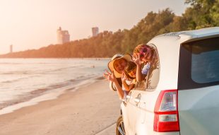 Happy family young woman with her daughter and son sitting in white car with hand up and look out from windows on the beach. Family holiday travel concept.